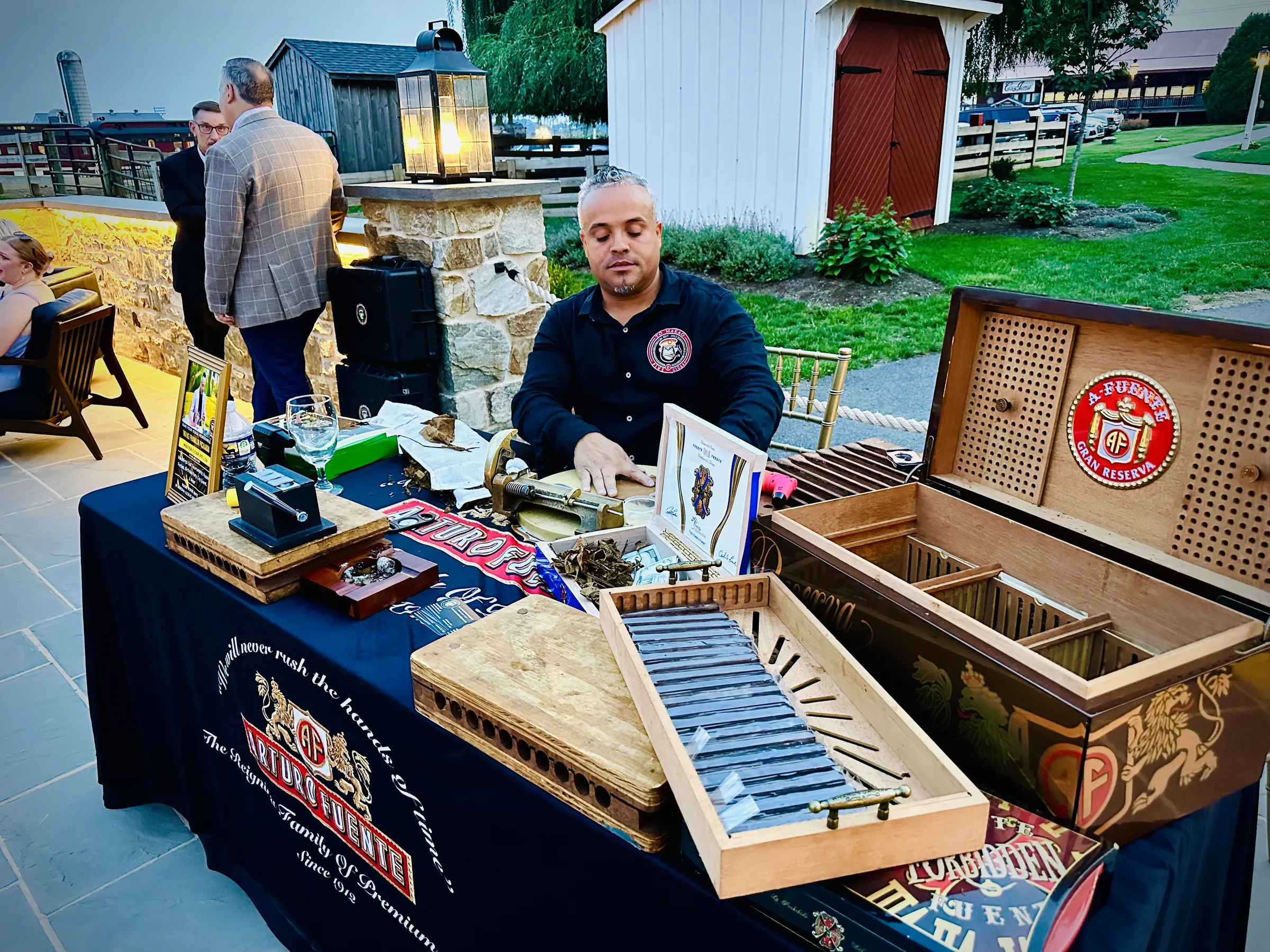 Franklin rolling at his table on a stone terrace at dusk, an open humidor beside him and guests on the lawn behind