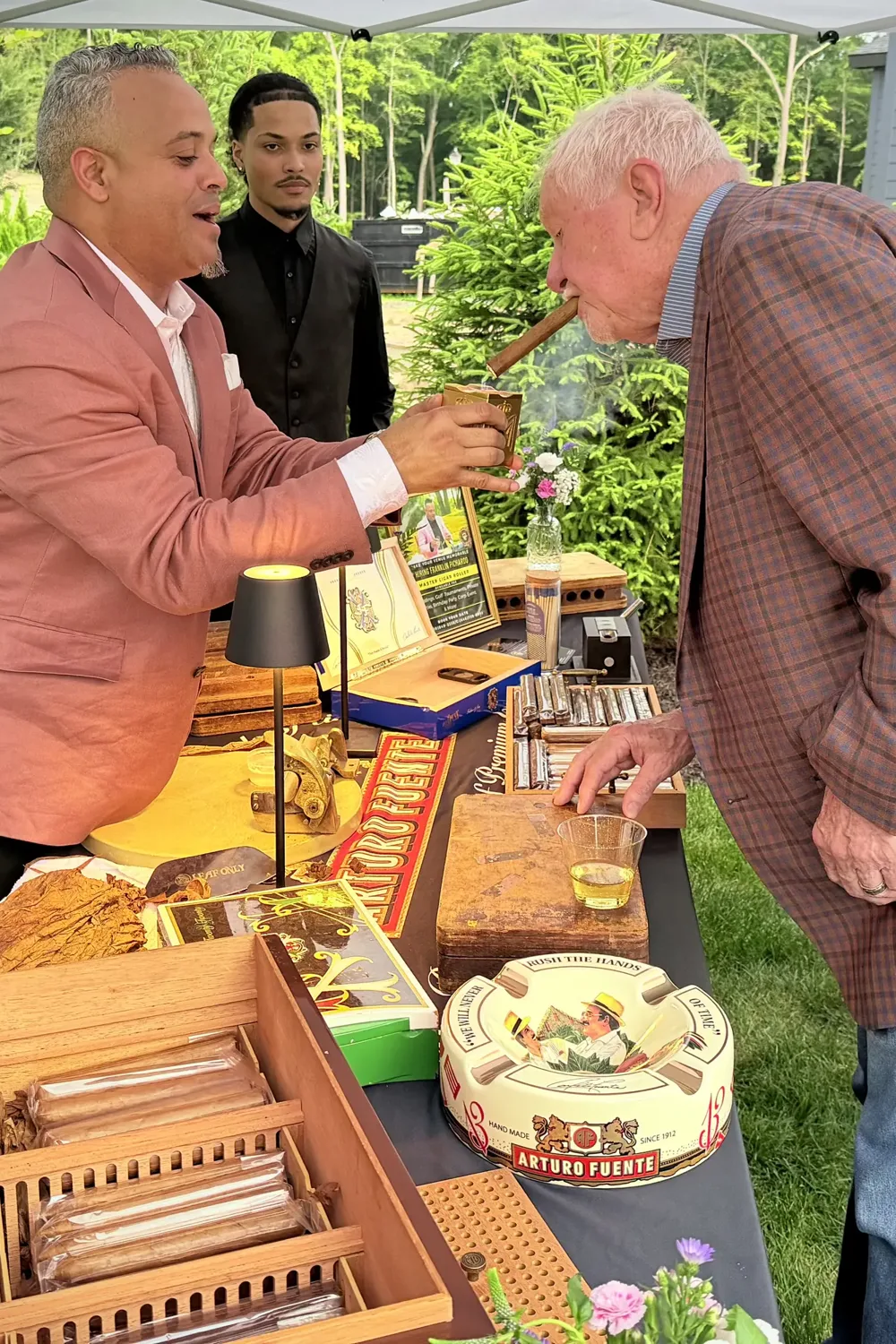 Franklin at his table at a garden party, an older guest leaning in to watch