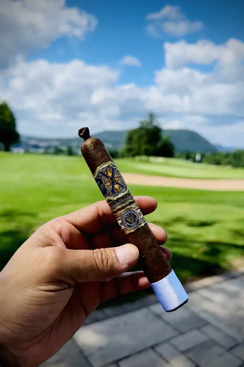 A finished cigar held up over a club terrace, the fairway and the hills behind