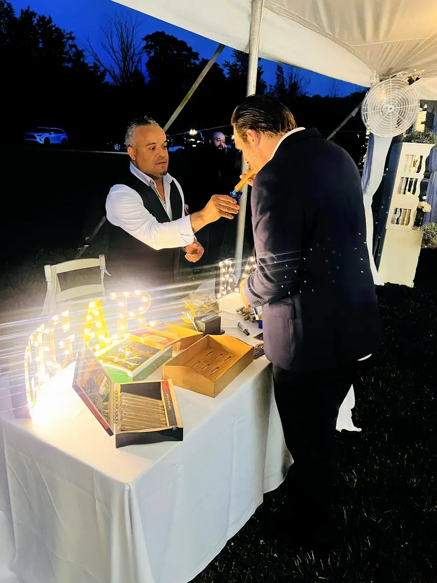 Franklin at his table under a white tent, handing a finished cigar to a guest
