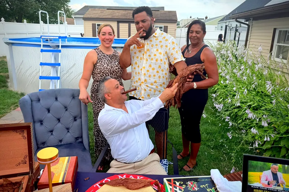 Franklin lighting a cigar at his table in a backyard, two guests laughing behind him