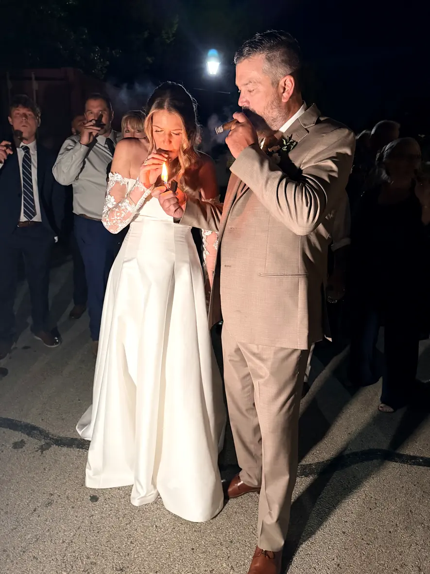 The bride drawing on a fresh cigar beside the groom, guests behind them