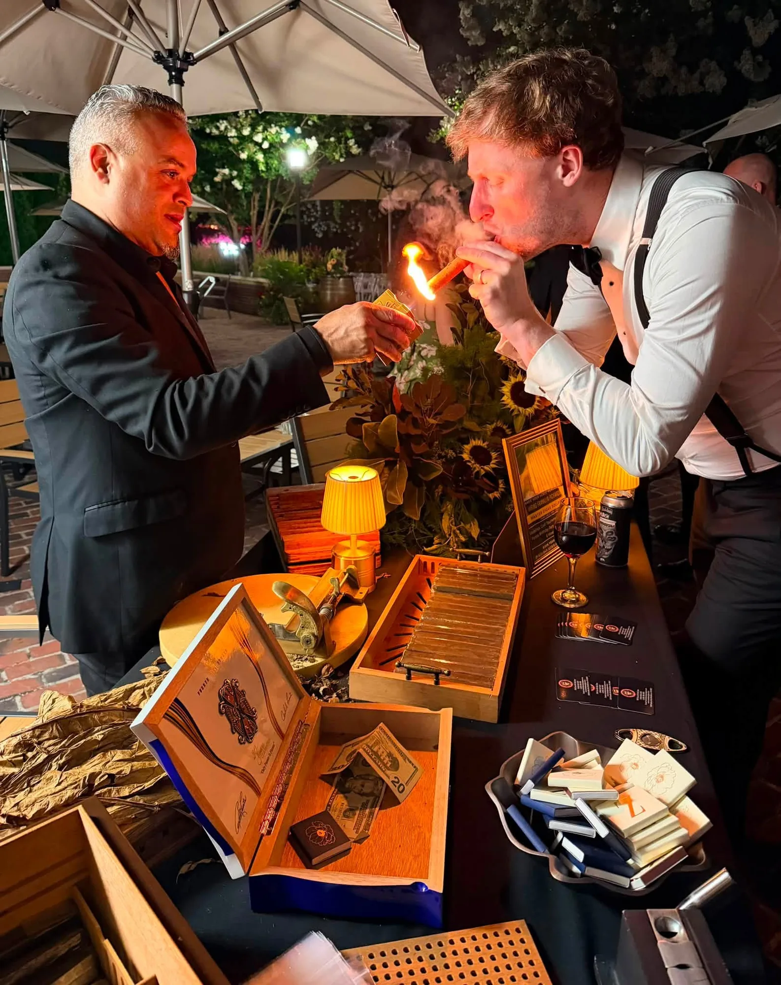 Franklin Pichardo lighting a guest’s cigar at his table after dinner, with the reception behind them