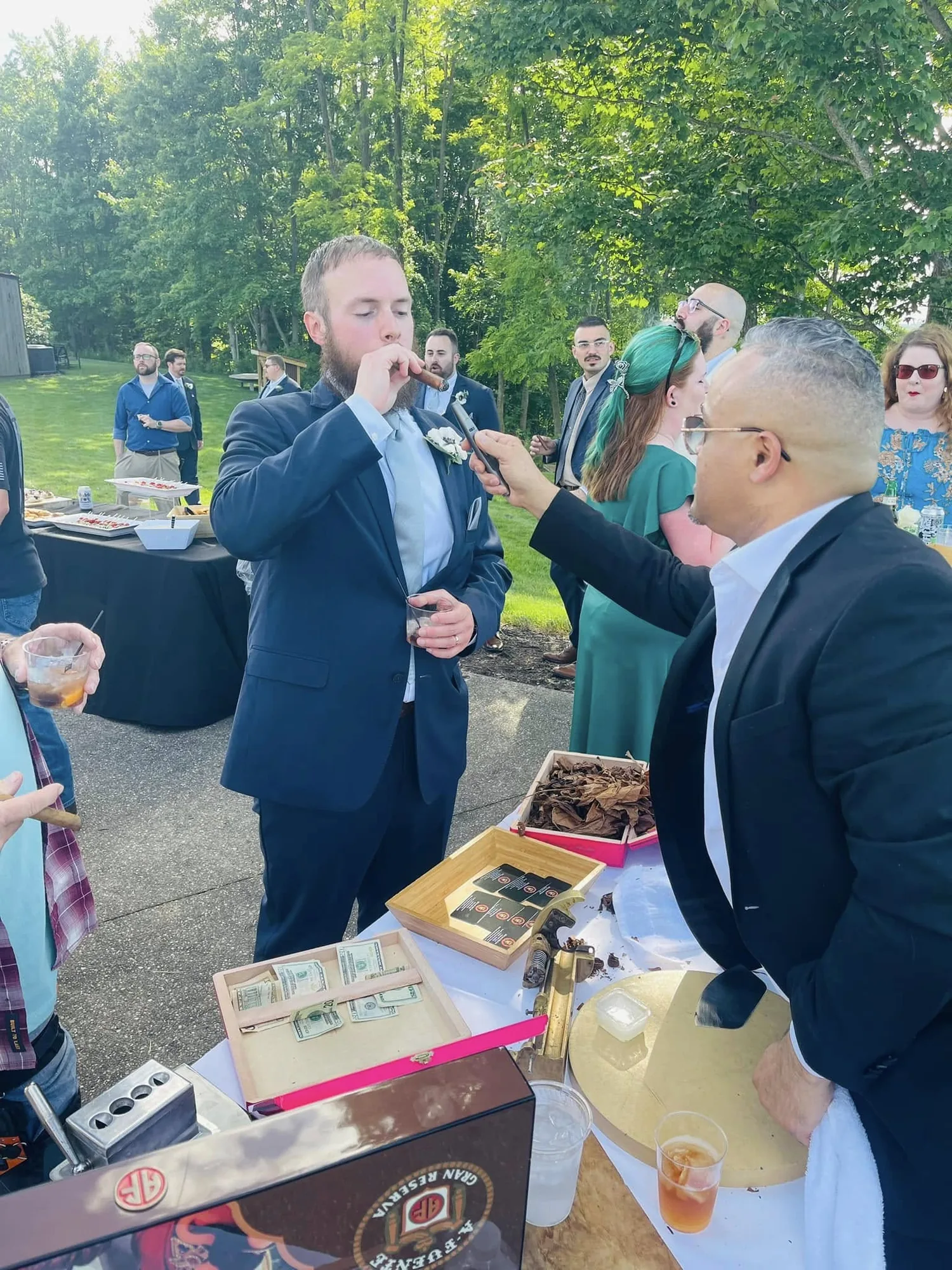 Franklin Pichardo lighting a guest’s cigar at an outdoor wedding reception, with guests on the lawn behind them