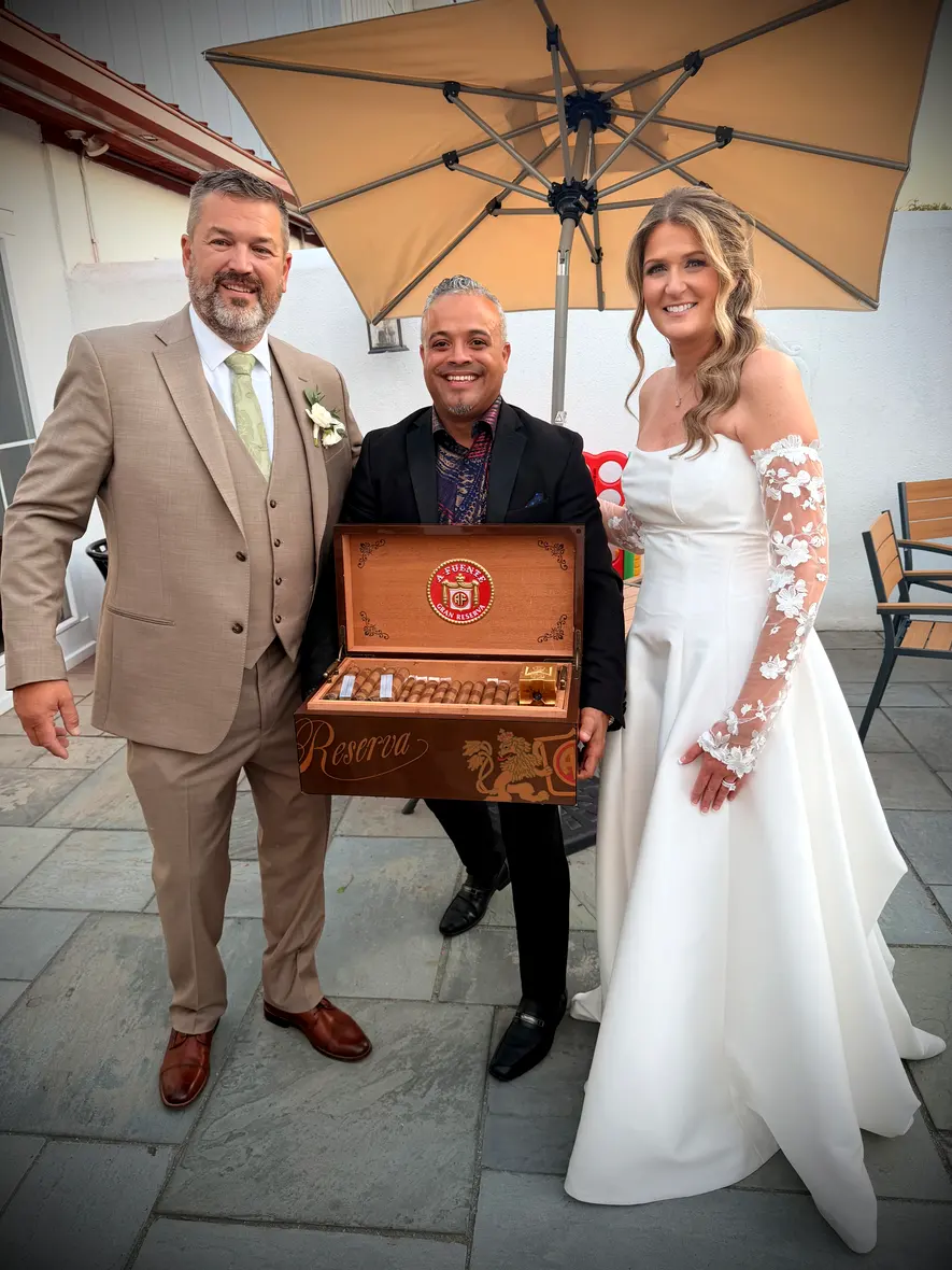 Franklin Pichardo with the bride and groom in a walled courtyard at dusk, holding his humidor open between them