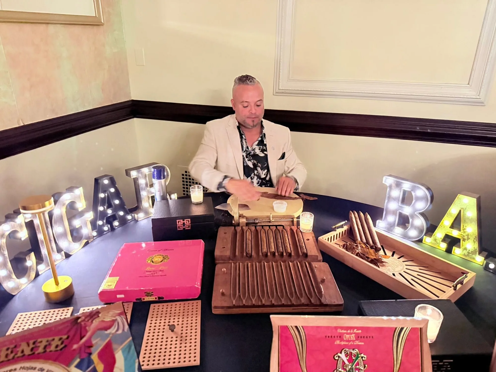 Franklin Pichardo seated at a cigar bar set up inside a wedding venue in New York, with lit-up letters and open boxes of cigars on a black tablecloth