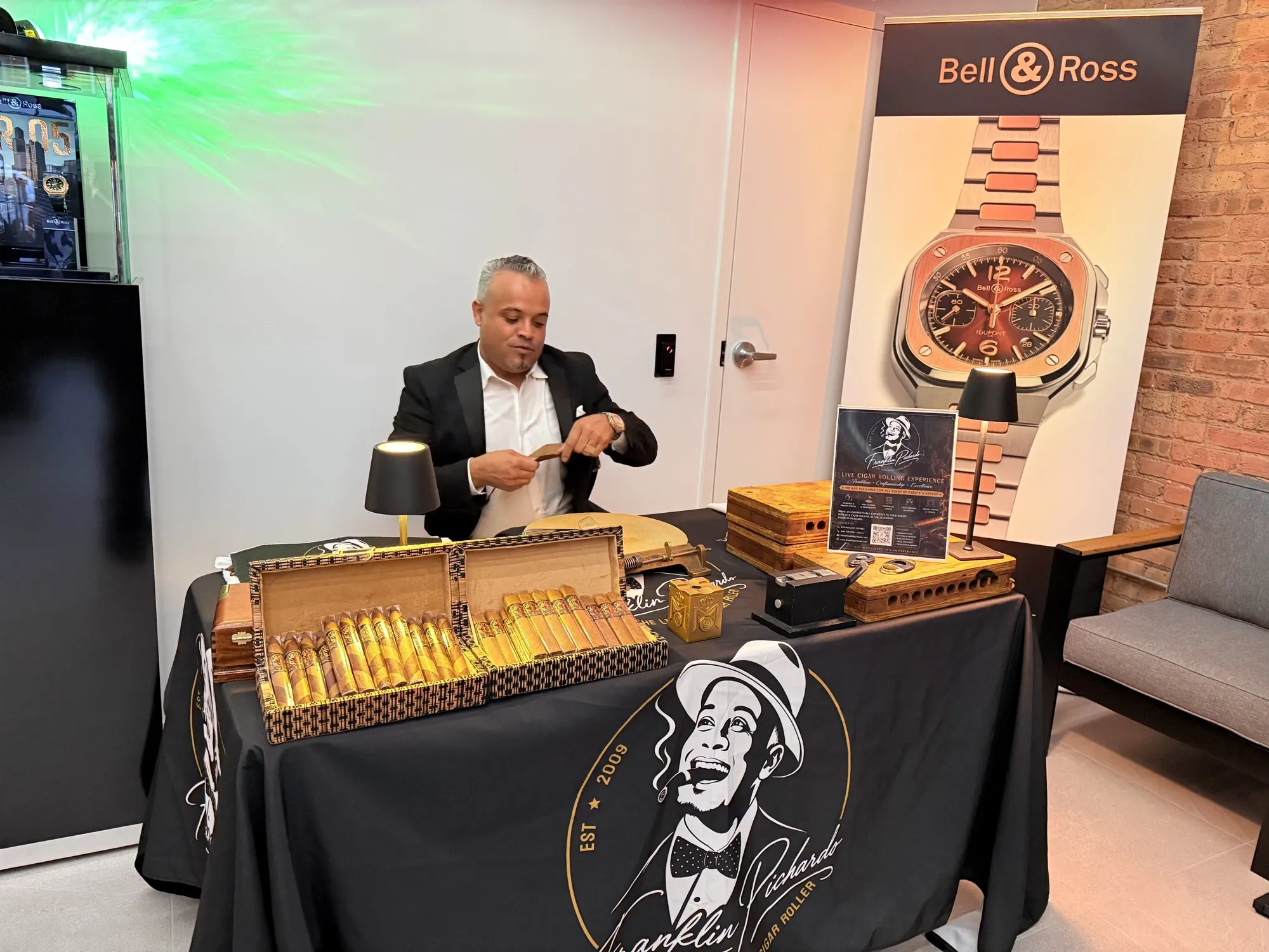 Franklin Pichardo rolling at his table beneath a tall Bell & Ross watch banner, with open boxes of finished cigars in front of him