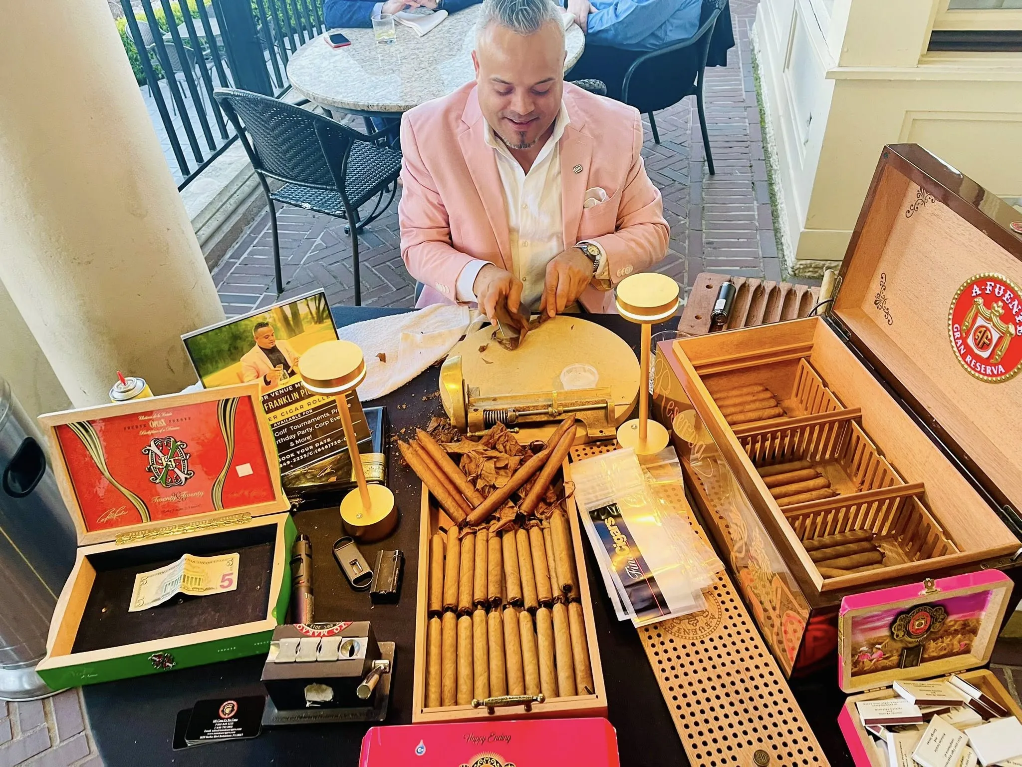 Franklin in a pink jacket rolling at his table on a terrace, boxes of cigars open around him and guests at the tables behind